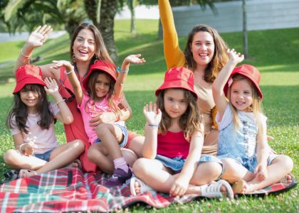 Two women and four girls enjoy a sunny picnic in Portugal, waving happily in the park.